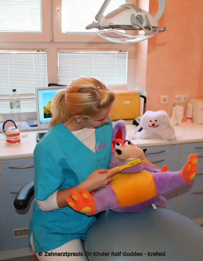 Mitarbeiterin zeigt einem Kind mit einer Handpuppe spielerisch das Zähneputzen in der Kinderzahnarztpraxis
